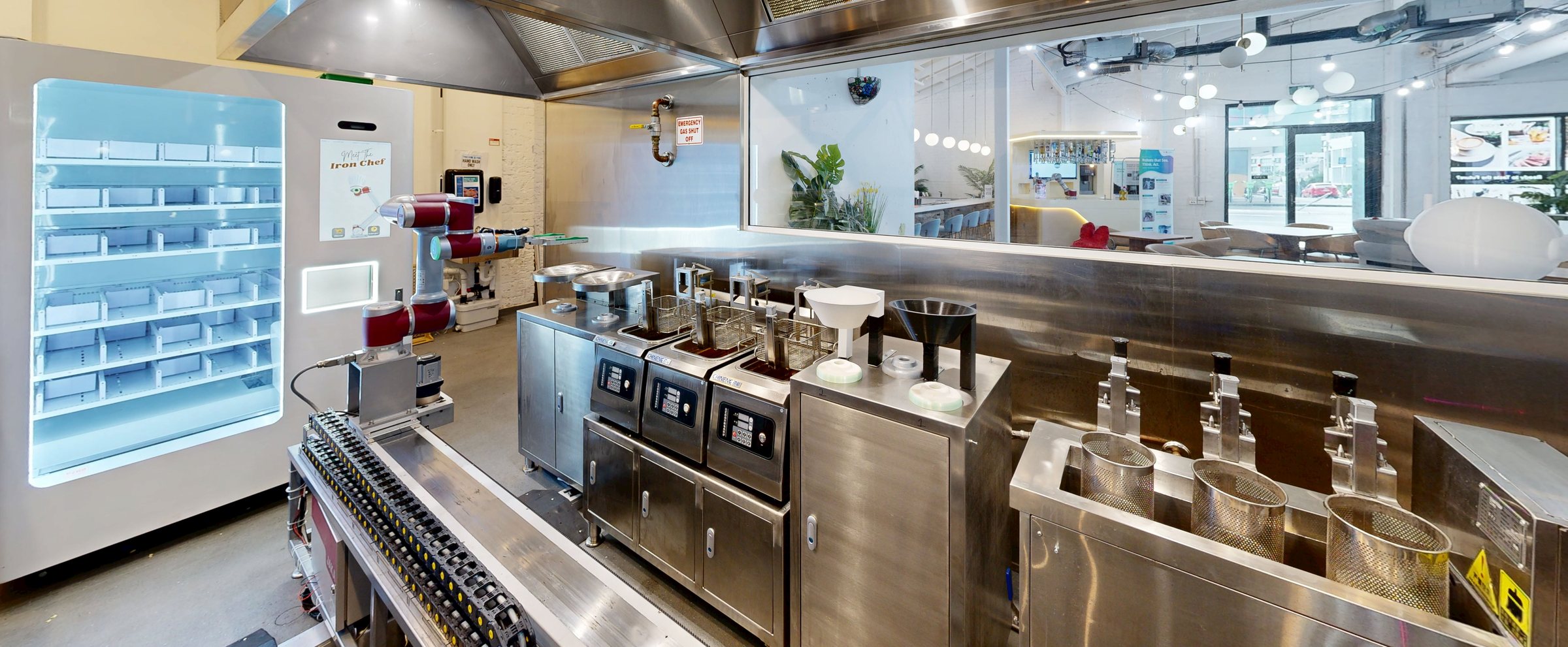 The kitchen line at 365 St Pauls Terrace in service, seen along its length: ingredient store, rail cobot, fryers, noodle baths, the pass