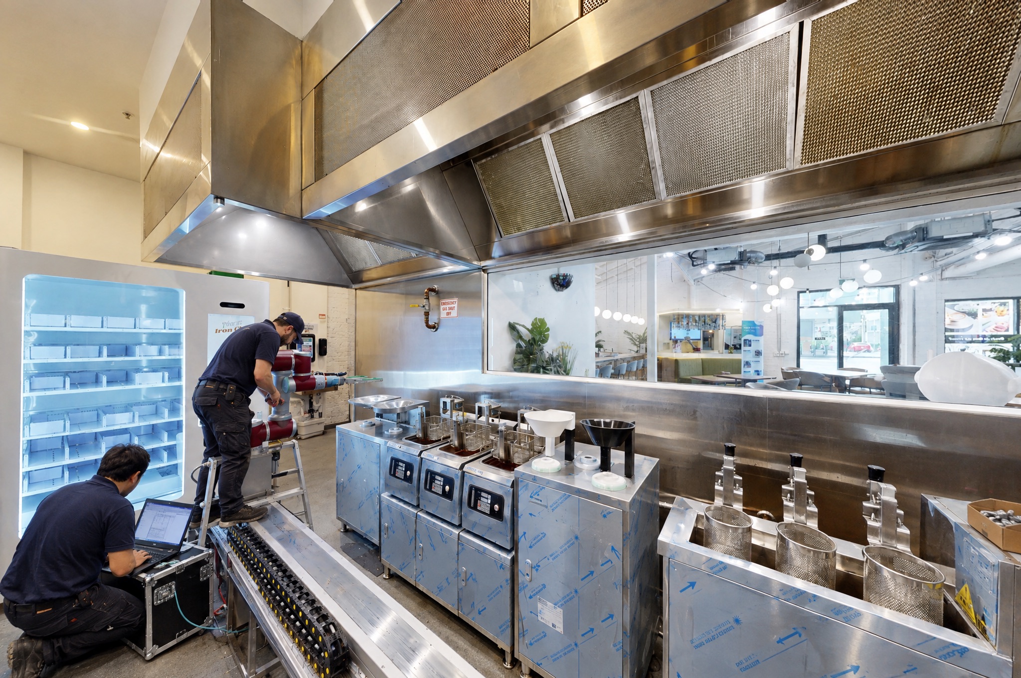 The same line on commissioning day: fryers and noodle baths still in blue protective film, a technician on a stepladder bolting the cobot to its rail, a second technician at a laptop on a flight case on the floor